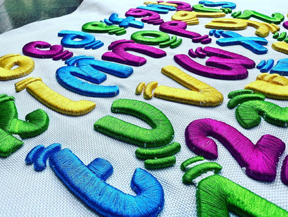 Wide close-up of colorful 3D puff embroidered alphabet letters and numbers spread across white fabric, stitched in bright blue, green, purple, and yellow thread.