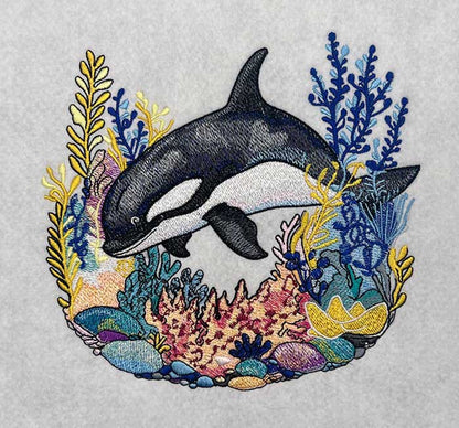 Embroidered design of an orca surrounded by colorful coral and seaweed on a light gray background.