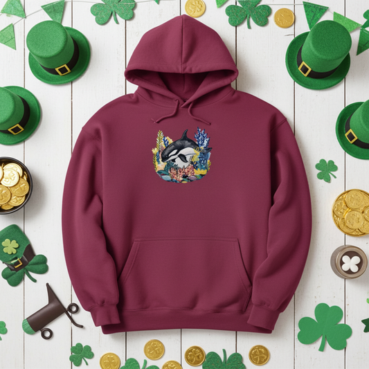 Burgundy premium hoodie with Playful Orca Coral embroidery showing an orca above colorful coral and sea plants, styled on a wooden background with shamrock decorations, green hats, and gold coins, original EmbroMania artwork.