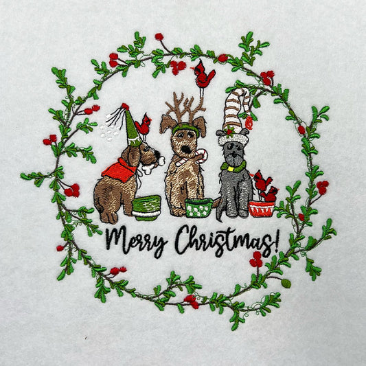 Close-up of embroidered Christmas wreath with three festive dogs and red cardinals, above the text “Merry Christmas!” on white fabric.