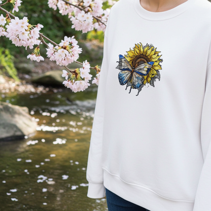 White sweatshirt worn outdoors near a flowing stream and blooming cherry blossoms, featuring detailed blue butterfly and sunflower embroidery on the chest