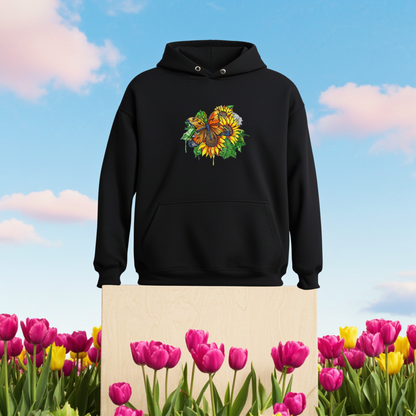 Black premium hoodie with Radiant Sunflower Butterfly embroidery featuring an orange butterfly over bright yellow sunflowers and green leaves, styled outdoors on a wooden block in a tulip field under a blue sky, original EmbroMania artwork.