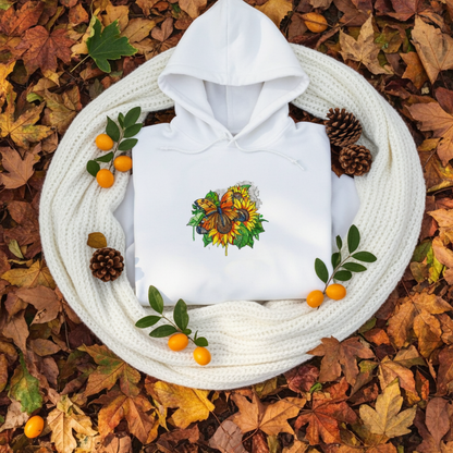 White premium hoodie with Radiant Sunflower Butterfly embroidery featuring an orange butterfly over bright yellow sunflowers and green leaves, folded flat on a white knit blanket surrounded by autumn leaves, pinecones, and small orange berries, original EmbroMania artwork.