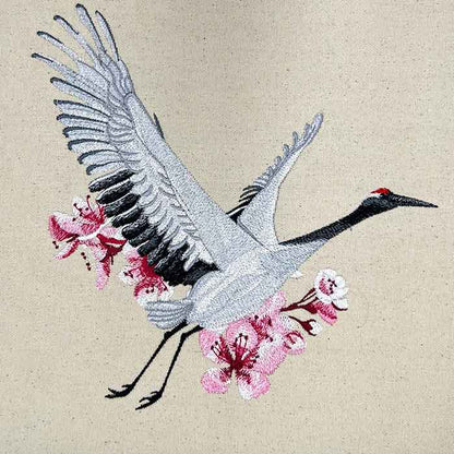 Embroidered white crane in flight with black wing accents and pink sakura cherry blossoms, elegant Japanese-inspired design, original EmbroMania artwork.