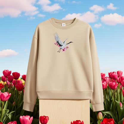 Beige unisex crewneck sweatshirt with sakura crane embroidery design, displayed on stand with tulip field and blue sky background.