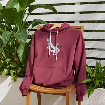 Burgundy premium hoodie with Sakura Crane embroidery showing a white crane in flight with pink cherry blossoms, styled on a wooden chair outdoors with lush green plants, original EmbroMania artwork.