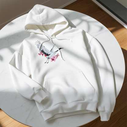 White premium hoodie with Sakura Crane embroidery showing a white crane in flight with pink cherry blossoms, laid flat on a round white table in natural light, original EmbroMania artwork.