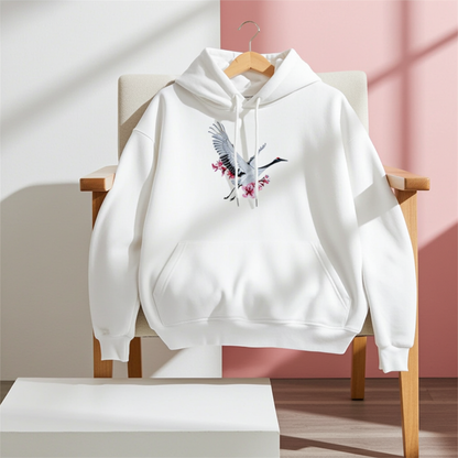 White premium hoodie with Sakura Crane embroidery showing a white crane in flight with pink cherry blossoms, hanging on a chair in a bright room with a soft pink wall, original EmbroMania artwork.