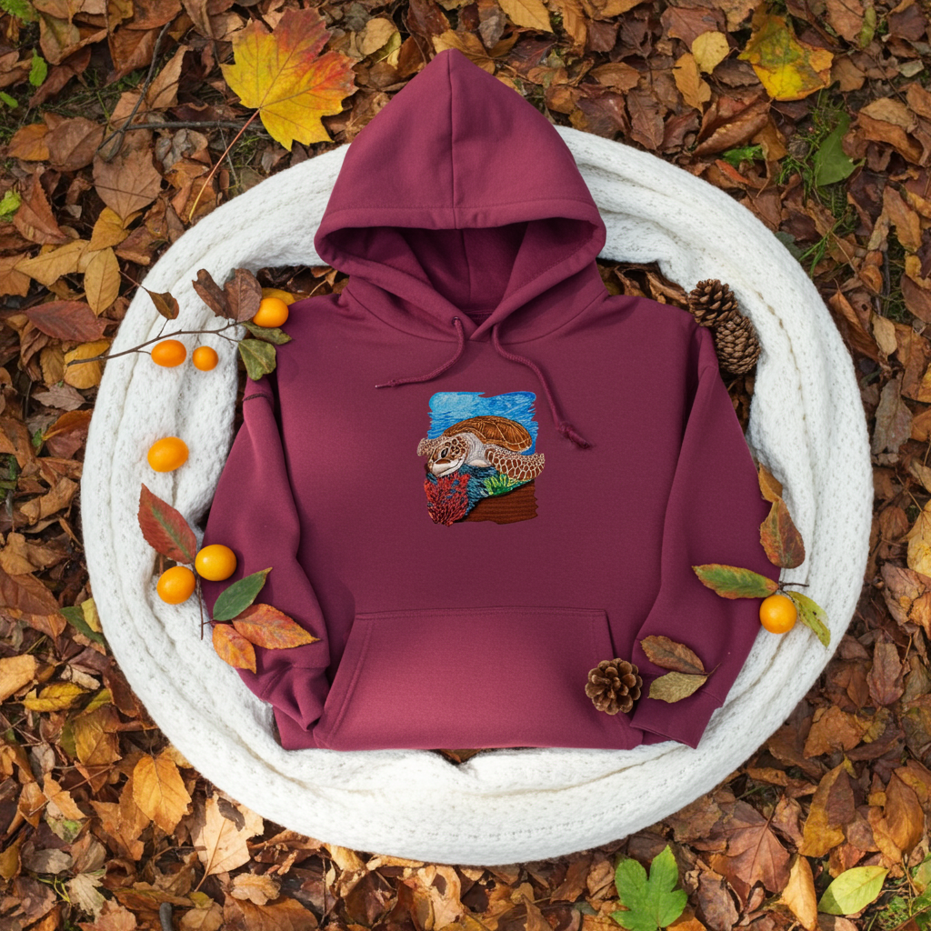Burgundy premium hoodie with Sea Turtle & Coral Reef embroidery showing a sea turtle above red coral and green sea plants on a blue ocean background, flat lay on a white knit blanket with autumn leaves, pinecones, and small orange berries, original EmbroMania artwork.