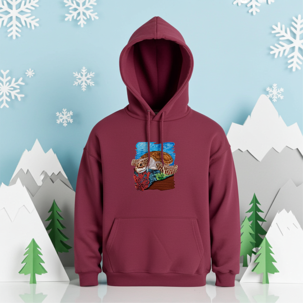 Burgundy premium hoodie with Sea Turtle & Coral Reef embroidery showing a sea turtle above red coral and green sea plants on a blue ocean background, styled in a winter scene with snowflakes, mountains, and pine trees, original EmbroMania artwork.