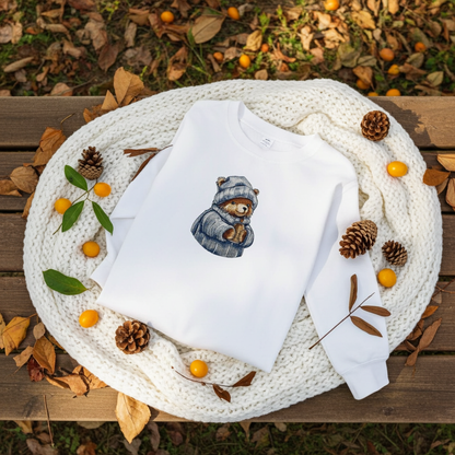 White sweatshirt laid on a chunky knit blanket outdoors, featuring a cute bear embroidery wearing a cozy hat and scarf, surrounded by autumn leaves, pinecones, and small orange berries