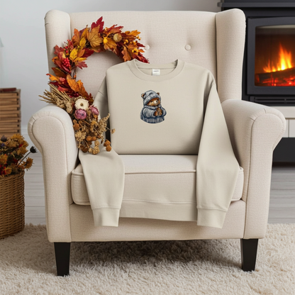 Mint green sweatshirt folded on a wooden stump, featuring a cute bear embroidery wearing a knit hat and sweater, styled with dried flowers and cotton stems on a warm neutral background