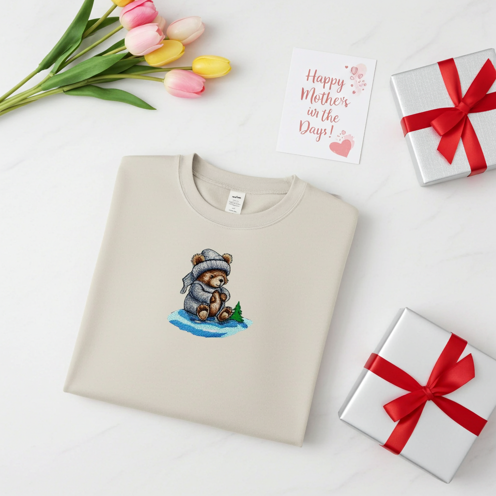 Mint green sweatshirt folded on a wooden stump, featuring a cute bear embroidery wearing a knit hat and sweater, styled with dried flowers and cotton stems on a warm neutral background