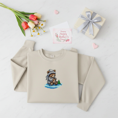 Mint green sweatshirt folded on a wooden stump, featuring a cute bear embroidery wearing a knit hat and sweater, styled with dried flowers and cotton stems on a warm neutral background