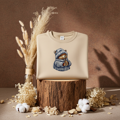 Soft beige sweatshirt folded on a wooden stump, featuring a cute bear embroidery wearing a knit hat and sweater, styled with dried flowers and cotton stems on a warm neutral background
