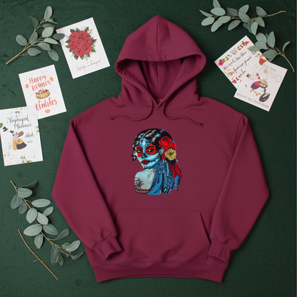 Burgundy premium hoodie with Sugar Skull Lady embroidery featuring a Día de los Muertos portrait in blue and red tones with floral accents, flat lay on a dark green background with eucalyptus leaves and greeting cards, original EmbroMania artwork.