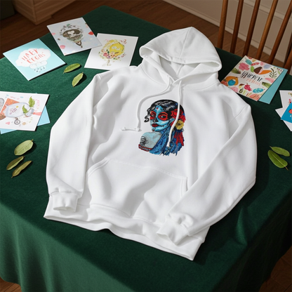 White premium hoodie with Sugar Skull Lady embroidery featuring a Día de los Muertos portrait in blue and red tones with floral accents, laid flat on a dark green surface with greeting cards and scattered leaves, original EmbroMania artwork.