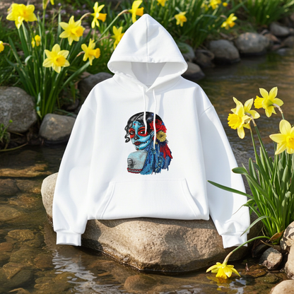 White premium hoodie with Sugar Skull Lady embroidery featuring a Día de los Muertos portrait in blue and red tones with floral accents, styled outdoors on a rock by a stream with yellow flowers, original EmbroMania artwork.