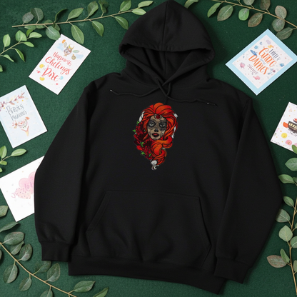 Black premium hoodie with Sugar Skull Rose Girl embroidery featuring a sugar skull portrait with bright orange hair and red roses, flat lay on a dark green background with greenery and greeting cards, original EmbroMania artwork.