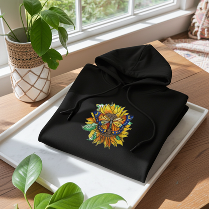 Black premium hoodie with Sun-Kissed Butterfly & Sunflower embroidery featuring a blue and orange butterfly on a golden sunflower with green leaves, folded flat on a white tray by a window with houseplants, original EmbroMania artwork.