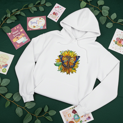 White premium hoodie with Sun-Kissed Butterfly & Sunflower embroidery featuring a blue and orange butterfly on a golden sunflower with green leaves, flat lay on a dark green background with eucalyptus leaves and greeting cards, original EmbroMania artwork.