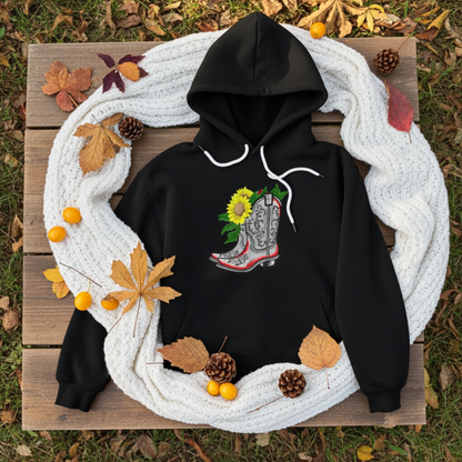 Black hoodie mockup with embroidered cowboy boot and sunflower appliqué on the chest, styled with autumn leaves and a cozy white knit blanket.
