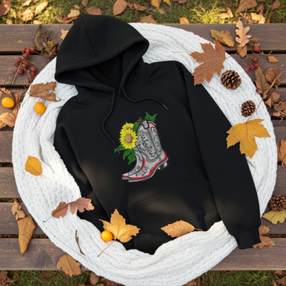 Black hoodie mockup with embroidered cowboy boot and sunflower appliqué on the chest, styled with autumn leaves and a cozy white knit blanket.
