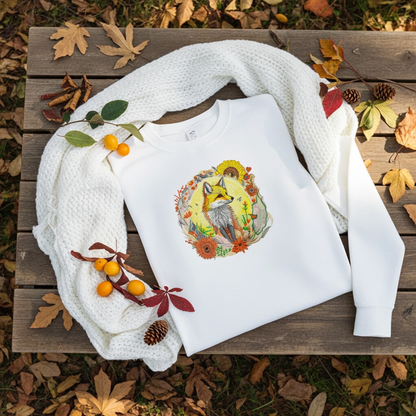 White unisex crewneck sweatshirt with sunlit fox and floral embroidery design, flat lay with autumn leaves, scarf, pinecones and berries.