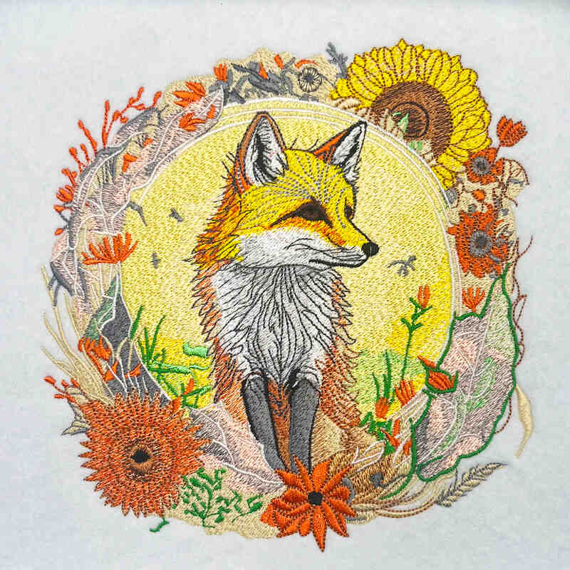 Embroidered fox portrait framed by sunflowers and wildflowers in warm golden, orange, and green tones, nature-inspired woodland design, original EmbroMania artwork.