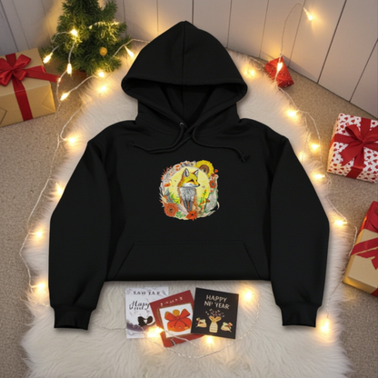 Black premium hoodie with Sunshine Fox & Sunflower embroidery featuring a fox framed by sunflowers and wildflowers in warm tones, styled on a cozy rug with string lights, greeting cards, and wrapped gifts, original EmbroMania artwork.