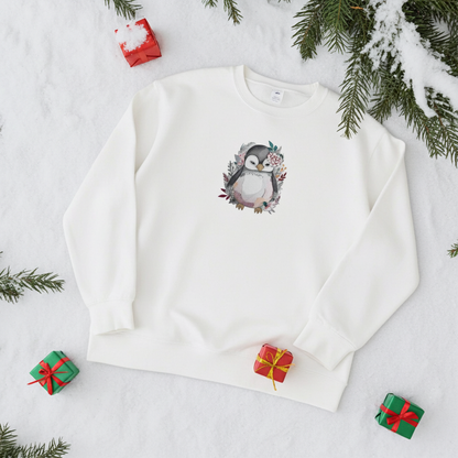 White sweatshirt laid flat on snow, featuring a cute floral penguin embroidery on the chest, styled with evergreen branches and small wrapped gift boxes around it