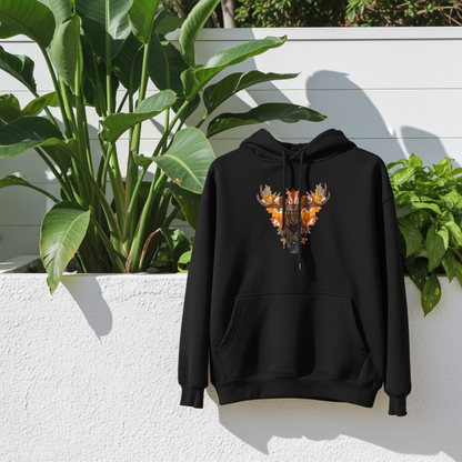 Black premium hoodie with Tribal Fire Moose embroidery featuring a moose head with bold antlers and tribal details in vibrant orange and gold tones, displayed against a white textured wall with lush green potted plants and natural sunlight, original EmbroMania artwork.