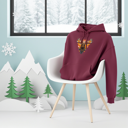 Burgundy premium hoodie with Tribal Fire Moose embroidery featuring a moose head with bold antlers and tribal details in vibrant orange and gold tones, displayed on a white modern chair against a winter landscape backdrop with snow-covered mountains, evergreen trees, and snowflakes, original EmbroMania artwork.