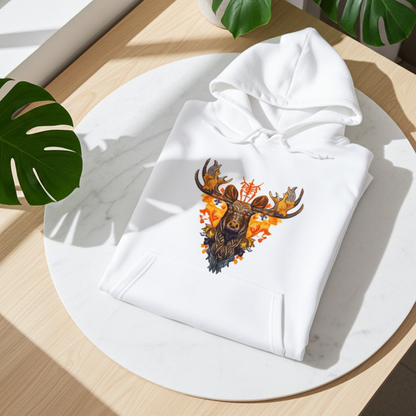 White premium hoodie with Tribal Fire Moose embroidery featuring a moose head with bold antlers and tribal details in warm orange and brown tones, folded on a round white table in bright natural light with tropical leaves, original EmbroMania artwork.