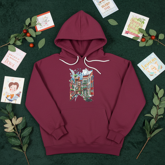 Burgundy premium hoodie featuring Urban Alley embroidery with a colorful cityscape sketch of buildings, windows, balconies, and plants in red, teal, green, and brown tones, styled flat-lay on a deep green background with greeting cards, leafy branches, and red berries, original EmbroMania artwork.