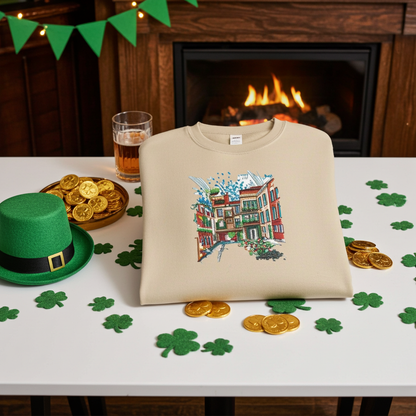 Beige unisex crewneck sweatshirt with urban cityscape embroidery design, folded on table with St Patrick’s Day decor, shamrocks, coins and fireplace background.