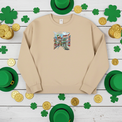Beige unisex crewneck sweatshirt with urban cityscape embroidery design, folded on table with St Patrick’s Day decor, shamrocks, coins and fireplace background.