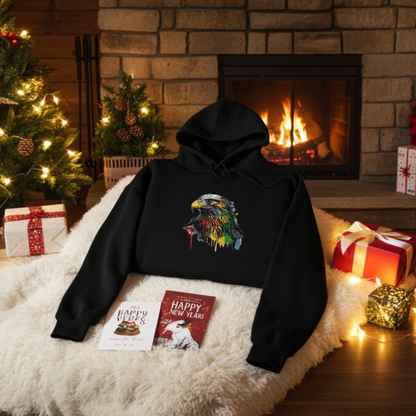 
Black hoodie featuring a vivid eagle head embroidery design with a multicolor paint-splash effect and drip accents on the chest, styled on a fluffy rug in a cozy holiday living room with a lit fireplace, Christmas tree, and gift boxes, original EmbroMania artwork.