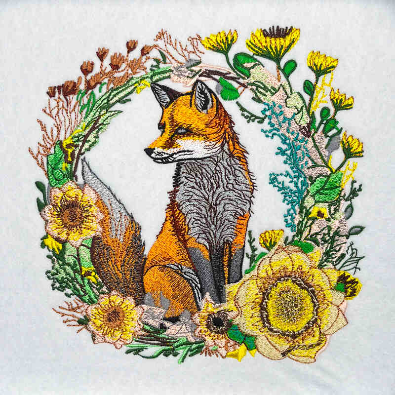 Wild fox and floral embroidery design featuring an orange fox with detailed fur stitching surrounded by a circular wreath of wildflowers and greenery in yellow, green, and brown thread, on a light background, original EmbroMania artwork.