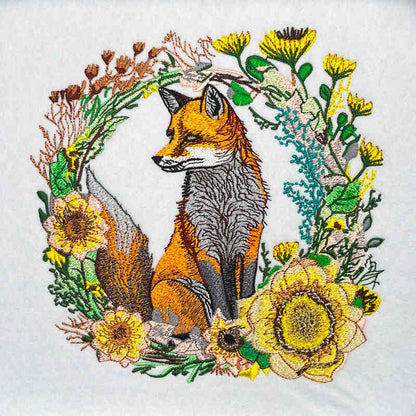 Wild fox and floral embroidery design featuring an orange fox with detailed fur stitching surrounded by a circular wreath of wildflowers and greenery in yellow, green, and brown thread, on a light background, original EmbroMania artwork.