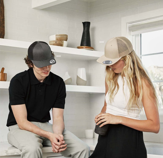 Two people wearing YP Classics trucker and snapback caps in a bright room; ideal base for custom 3D puff embroidery by EmbroMania, Dubai.