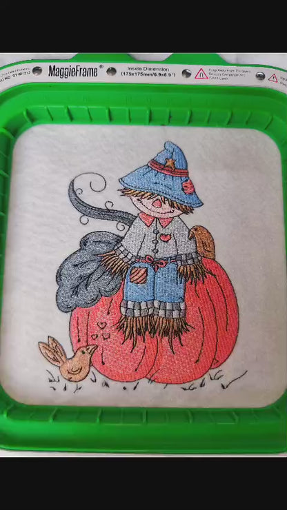 Embroidered fall design featuring a cute scarecrow with a blue hat and patchwork clothes sitting on a pumpkin, surrounded by birds and autumn leaves. Perfect for hoodies, sweatshirts, and tote bags.