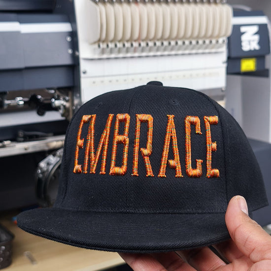 Close-up of a black cap featuring “EMBRACE” in Metro-style multicolor 3D puff embroidery, shown during stitching on an embroidery machine, ending with the Embromania logo and “Crafting Visions” text.